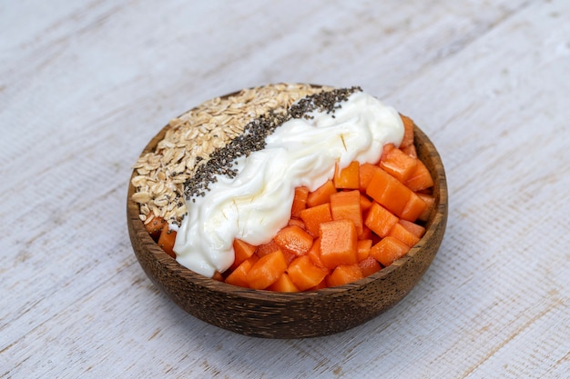 Slices of ripe sweet papaya fruit with oat flakes, chia seeds and white yogurt on coconut bowl on white wooden background, close up . The concept of healthy eating