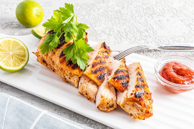 Slices of sliced grilled chicken fillet breast on a serving white rectangular plate with sauce and lime large species fork healthy dinner food