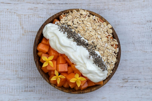 Slices of sweet papaya with oat flakes chia seeds and white yogurt in coconut bowl on white wooden background close up