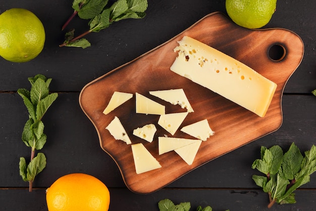 Slices of yellow hard cheese on a wooden board with citrus fruits with fresh mint on a wooden black background Closeup of a still life View from above