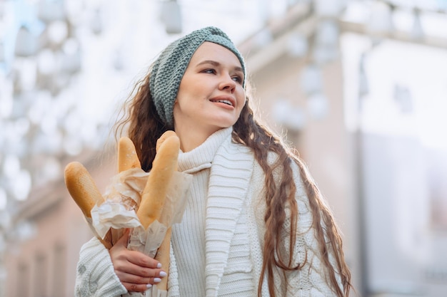 Slightly tilted portrait of young model posing for bakery advertising product. Photo on the street on blurry background. An all-white knit suit and crocheted headband. Holding baguettes in your hands.