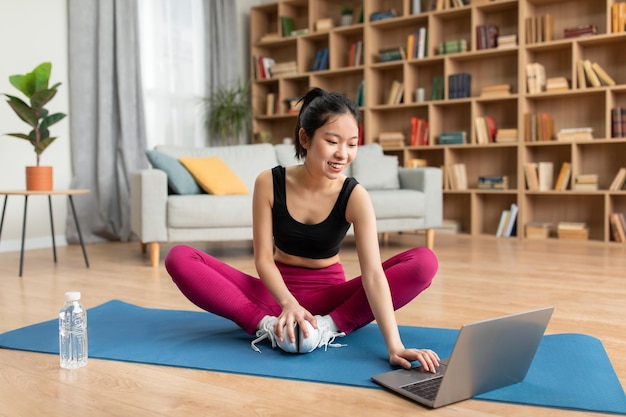 Slim asian lady choosing online tutorials on laptop for training at home sitting on yoga mat in