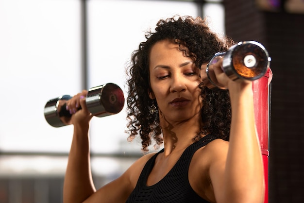 Slim athletic African American woman in sportswear doing exercises with dumbbells on a bench press in the gym copyspace