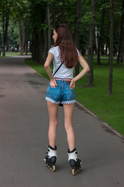 Slim brunette teen in denim shorts riding in the Park, rollerblading and keeps hands in her pockets