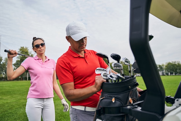 Slim dark-haired Caucasian female golfer in sunglasses standing behind her personal trainer in a cap