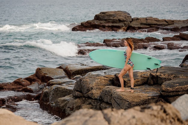 Slim figure girl in the multi colored swimsuit standing with the surfboard on the rocks on the beach of Atlantic ocean