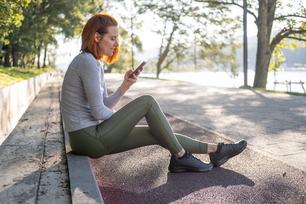 Slim ginger woman in sport clothes using mobile phone at park health wellness fitness lifestyle