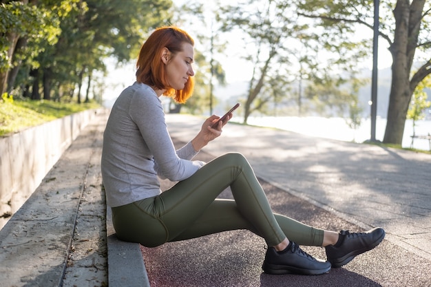 Slim ginger woman in sport clothes using mobile phone at park. Health, wellness, fitness lifestyle.