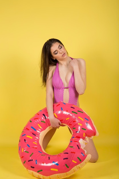 Slim girl in bikini with inflatable donut posing against yellow wall