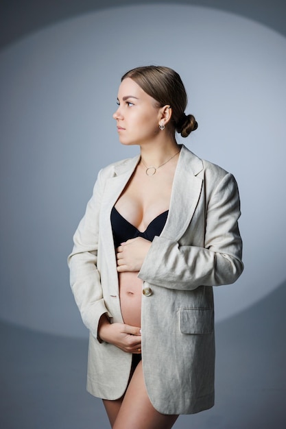 A slim pregnant woman in a long jacket and underwear stands in the studio on a white background holding her hand on her stomach Women's underwear for pregnant women