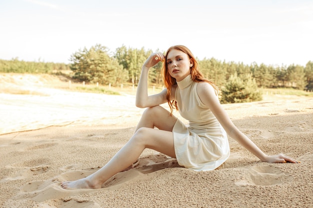 slim redhead woman in white dress sitting barefoot on sandy shore