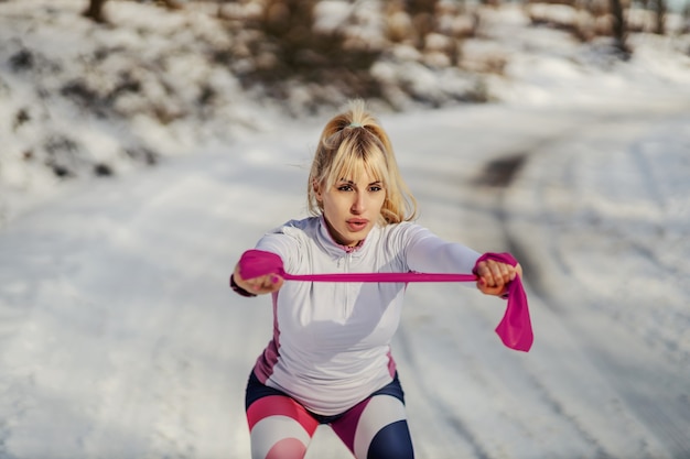 Slim sportswoman doing exercises with power rubber while standing in nature at snowy winter day.