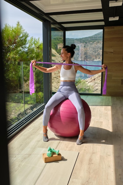 Slim sportswoman in sportswear sitting on fitness ball and doing exercise with rubber band during workout in studio in sunny day