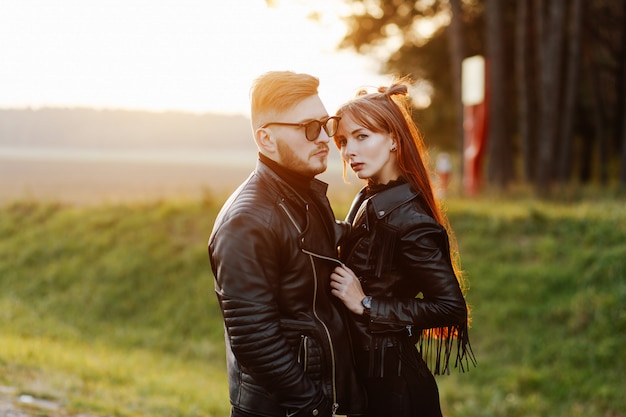 Slim stylish girl with long red hair hugging a brutal guy with a beard and wearing sunglasses at sunset