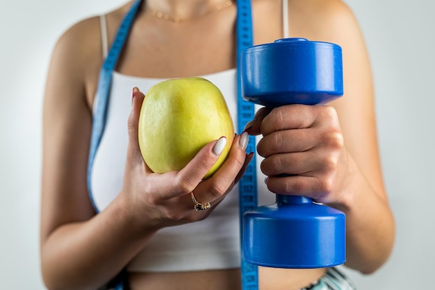 Slim unrecognizable woman with tape measure holding blue dumbbells and apple