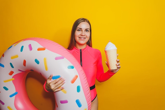 A slim woman in a bright pink swimsuit on a yellow background She is holding an inflatable pool donut Beach fashion