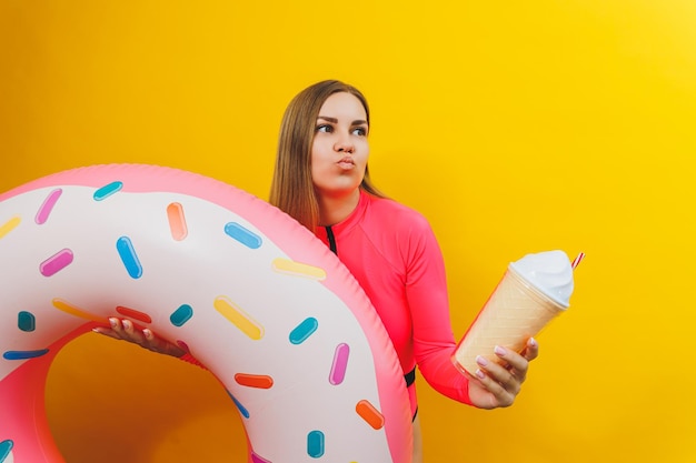 A slim woman in a bright pink swimsuit on a yellow background She is holding an inflatable pool donut Beach fashion