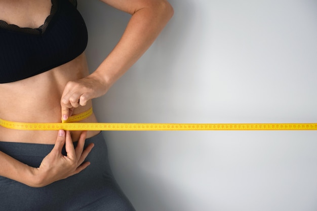 Slim woman is measuring her waist size with yellow tape measure on gray background.