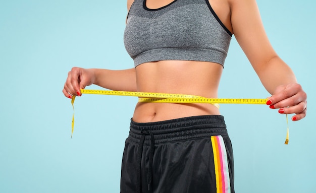 Slim woman measuring her waist's size with tape measure. Isolated on blue background.