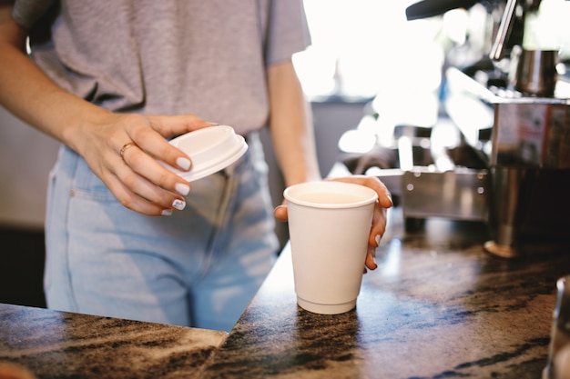 A slim young beautiful girl,dressed in casual outfit, gently puts the lid on the coffeein a cozy coffee shop. .