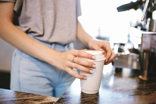 A slim young beautiful girl,dressed in casual outfit, gently puts the lid on the coffeein a cozy coffee shop. .