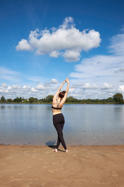 Slim young woman stretching on the sand near lake.