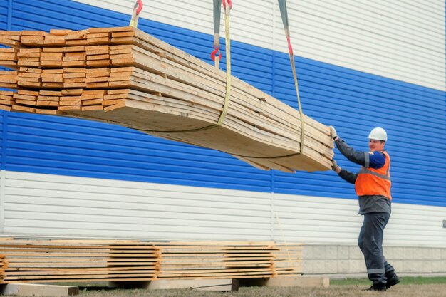 Slinger in helmet and signal vest unloads wooden planks outside on summer day