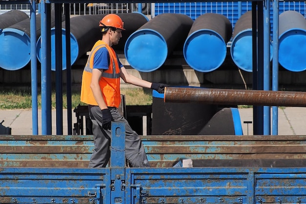 Slinger in helmet and vest unloads metal pipes outside on summer day Loading metal pipes for gasification