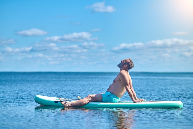 Slow life. Gray-haired bearded man sitting sideways to camera meditating on paddle board on water on sunny day