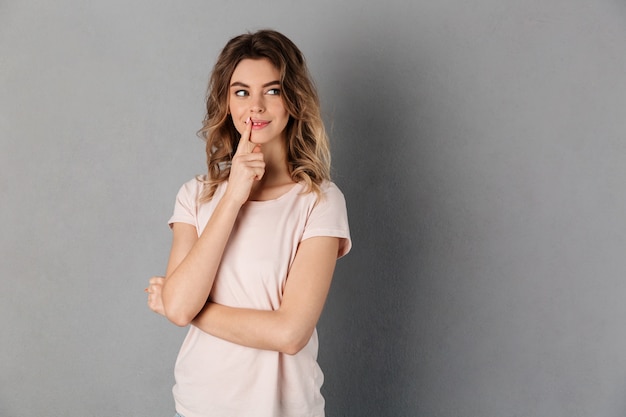 Sly pensive woman in t-shirt holding finger on mouth and looking away over grey