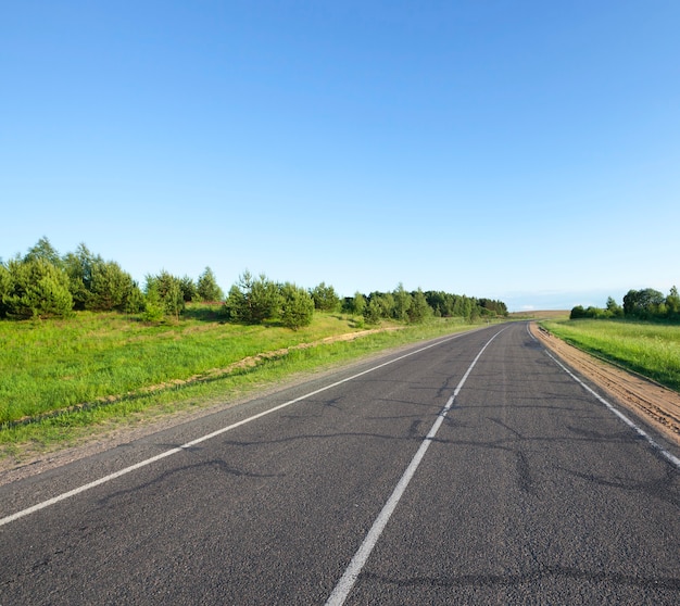 A small asphalt road in the summer. The roadway is divided by a white line of road markings for traffic in various directions. Blue sky and green young trees spruce, grass on a of a landscape