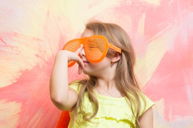 Small baby girl or cute child with adorable face and blonde hair in yellow shirt and orange sunglasses posing on colorful abstract background