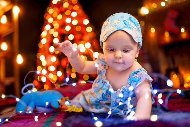 Small baby girl in pajama playing with garland while lying on checkered plaid near Christmas tree