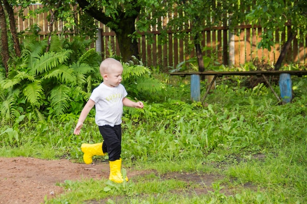 A small baldheaded boy in yellow boots runs in the countryside through puddles in the fresh air