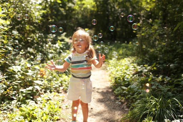 a small beautiful blonde girl in a light Tshirt and white shorts catches soap bubbles in the park