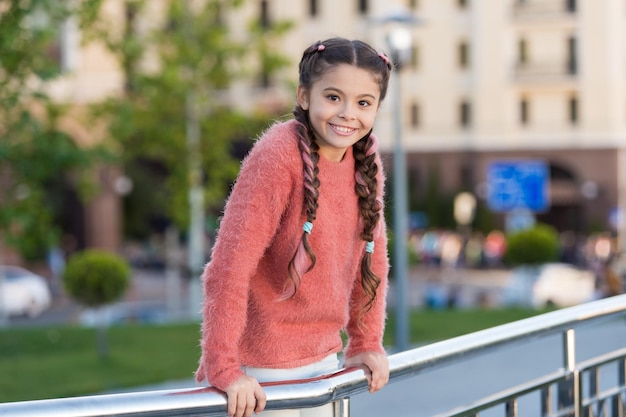 Small beauty Small child with brunette hair plaits smiling in casual fashion style Happy small girl with casual look on urban background Adorable small kid with lovely smile on summer day