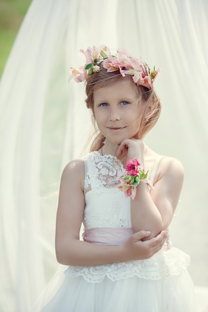 A small blonde European girl in a beautiful dress poses near the scenery in the field. Canopy, flowers, tenderness, wreath,