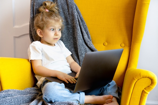 A small blonde girl looks very surprised and looks at her laptop while sitting in a chair at home