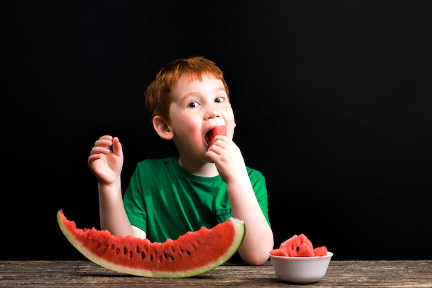 A small boy bites slices and eats chunks of red juicy watermelon sliced on the table