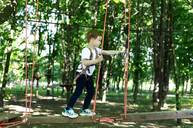 Small boy in climbing gear is walking along a rope road in an adventure Park holding on to a rope