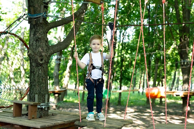 Small boy in climbing gear is walking along a rope road in an adventure Park holding on to a rope