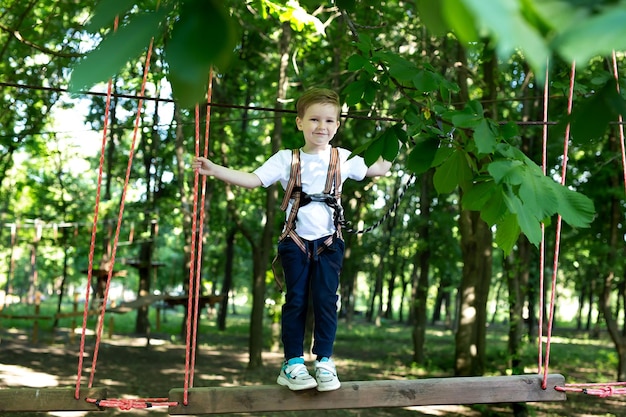 Small boy in climbing gear is walking along a rope road in an adventure Park holding on to a rope