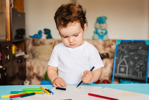 A small boy draws on sheets of paper lying on the table with colored pencils