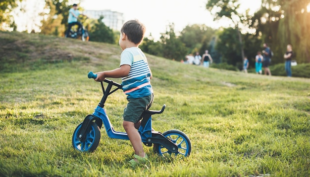 Small boy riding a bike in the park looking at other children walking with their parents