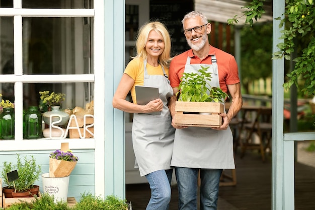 Small business concept smiling mature greenhouse owners couple posing near own cafe