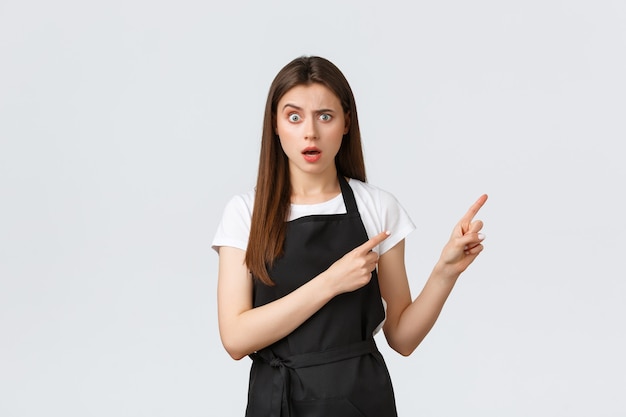 Small business, employees and coffee shop concept. Confused barista in black apron frowning and looking concerned as pointing upper right corner worried, standing white background