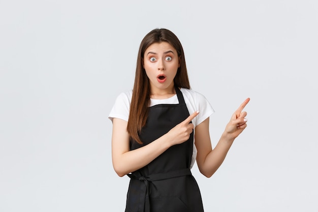 Small business, employees and coffee shop concept. Shocked and excited brunette female barista showing way at banner. Impressed grocery store saleswoman pointing upper right corner