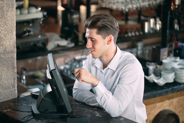 Small business, people and service concept - happy man or waiter in apron at counter with cashbox working at bar or coffee shop.