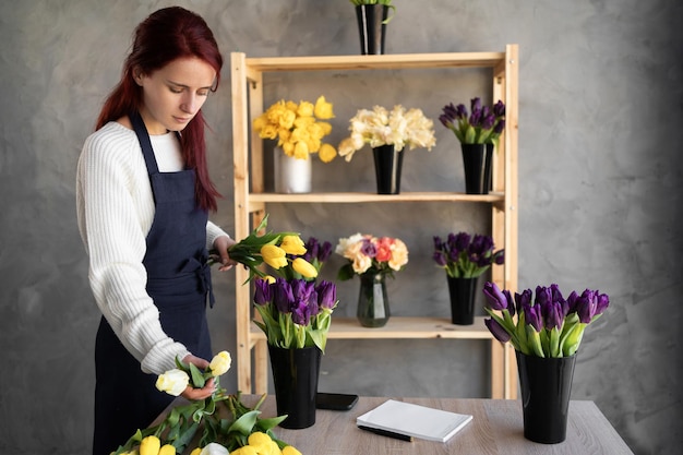 Photo small business a woman florist in a flower shop near a showcase with tulips in an apron collects a bouquet of tulip flowers flower delivery ordering