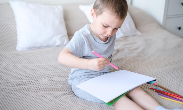 A small caucasian boy in a gray Tshirt and shorts on the bed draws with colored pencils in a sketch